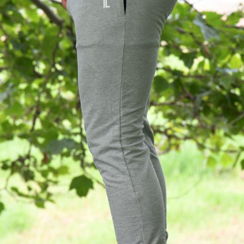 Outdoors on grass with leafy branches behind, a person wears grey Genesis Joggers featuring an adjustable fit and zipper pockets, paired with white slip-on shoes.