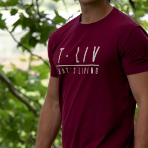 A man with long hair and sunglasses stands outdoors in front of leafy green trees, wearing the Genesis Tee - Maroon, a men’s performance t-shirt with tapered fit, scoop bottom, and "T. LIV THAT'S LIVING" printed on it.