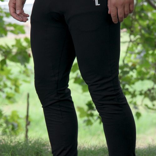 A person stands outdoors on grass wearing Genesis Joggers - Black and white slip-on shoes, with only the lower half of their body visible against a backdrop of green foliage and a blurred background.