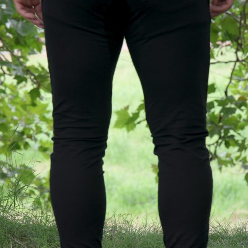 A person standing outdoors on grass, seen from behind, wearing Genesis Joggers - Black and white shoes. Green leaves and branches frame the upper part of the image.