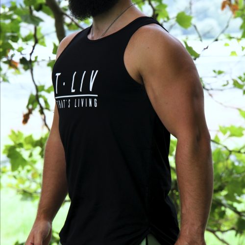 A bearded man in a black cap and Genesis Tank - Black with scoop hem, paired with light green athletic pants, stands outdoors before leafy trees, gazing to the side.