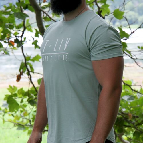 A bearded man stands outdoors near leafy trees, wearing the Genesis Tee - Mint Green with a tapered fit and scoop bottom, paired with black shorts. He looks off to the side; greenery and blurred water fill the background.