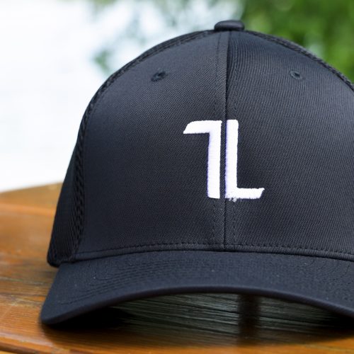 The Mesh Ball Cap - Black features a minimalist, breathable mesh design with flex fit and a white embroidered “7L” logo on the front. It’s displayed outdoors on a wooden surface with greenery in the background.