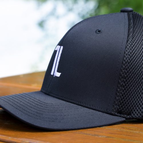 The Mesh Ball Cap - Black, featuring a white "TL" embroidered front, flex fit, and breathable mesh sides, rests on an outdoor wooden surface with its curved brim highlighted against blurred green foliage.