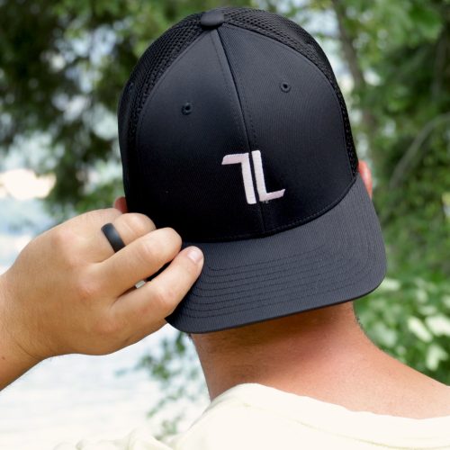 A person adjusts their Mesh Ball Cap - Black, featuring a white "7L" logo and a breathable flex fit design. The cap is viewed from behind against a serene backdrop of green foliage and blurred water.