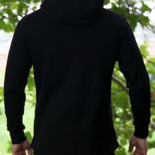 A person with light brown hair, wearing the black SPEKTR Hoodie, stands outdoors facing away, surrounded by green foliage.