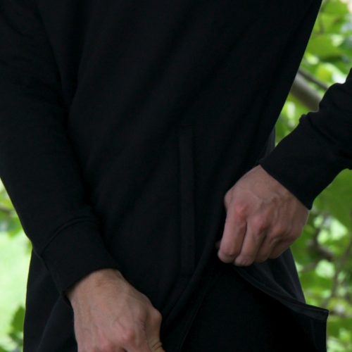 A person wearing the SPEKTR Hoodie-Black lifts its hem with one hand. Their face is hidden, and green leaves appear blurred in the background.