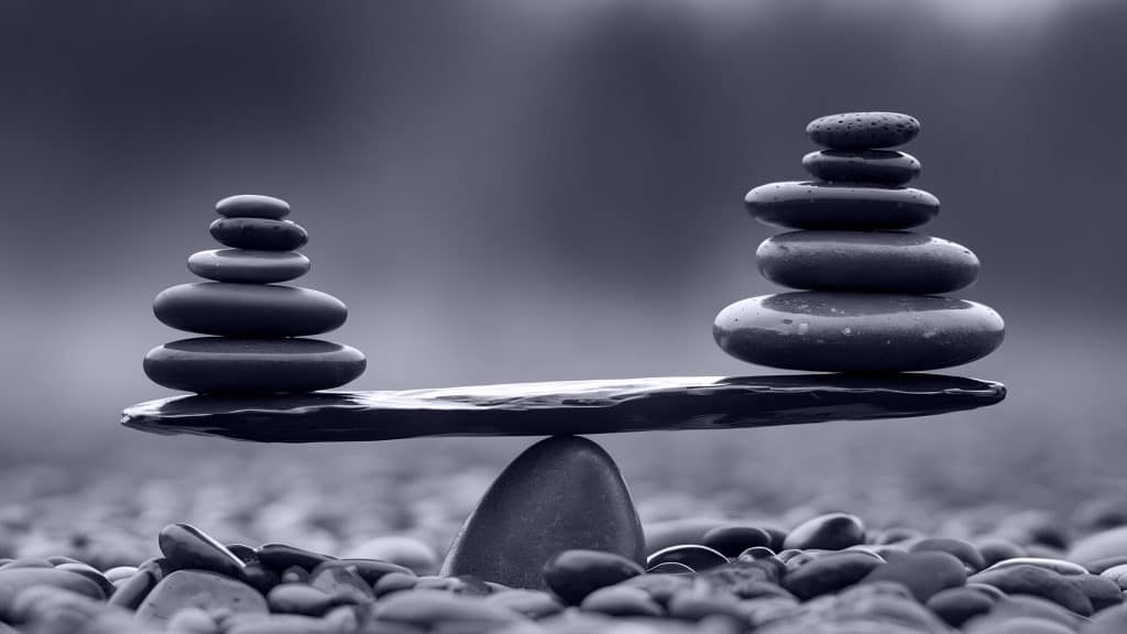A balancing stone seesaw with stacks of smooth pebbles on each side, set on a bed of small rocks. One stack is taller but both sides are balanced. The background is blurred, creating a calm, harmonious scene.