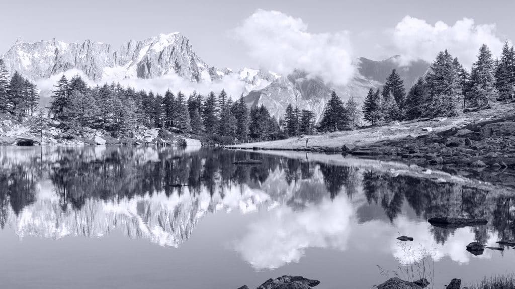 A serene mountain lake reflects pine trees and snow-capped peaks under a partly cloudy sky. Rocks and sparse grass line the shoreline, creating a peaceful natural landscape. The image is in black and white.
