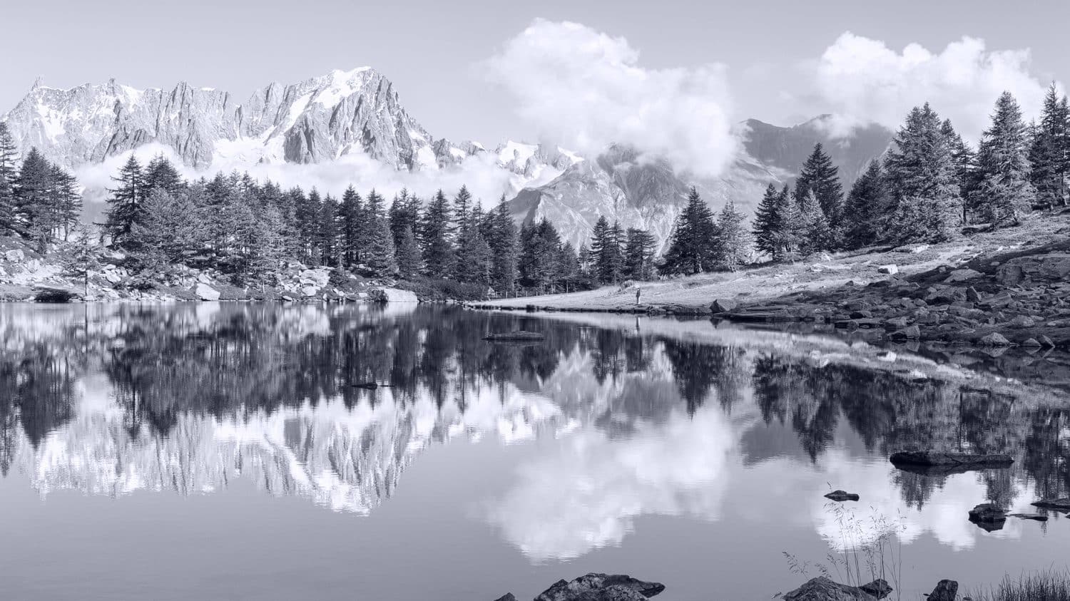 A serene mountain lake reflects pine trees and snow-capped peaks under a partly cloudy sky. Rocks and sparse grass line the shoreline, creating a peaceful natural landscape. The image is in black and white.