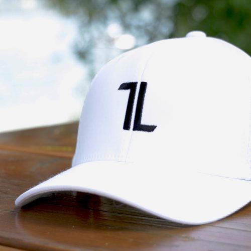 The Mesh Ball Cap - White is a minimalist, breathable flex fit cap with a black "1L" logo on the front, shown resting outdoors on wood with a blurred green and white background.