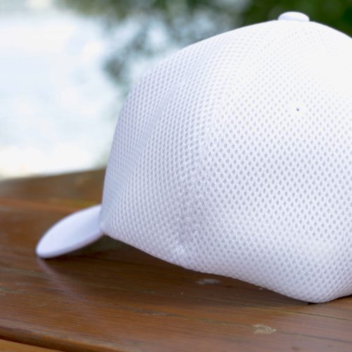 The Mesh Ball Cap - White, featuring a minimalist design with flex fit and breathable mesh, rests upside down on an outdoor wooden surface against a backdrop of blurred greenery and water.