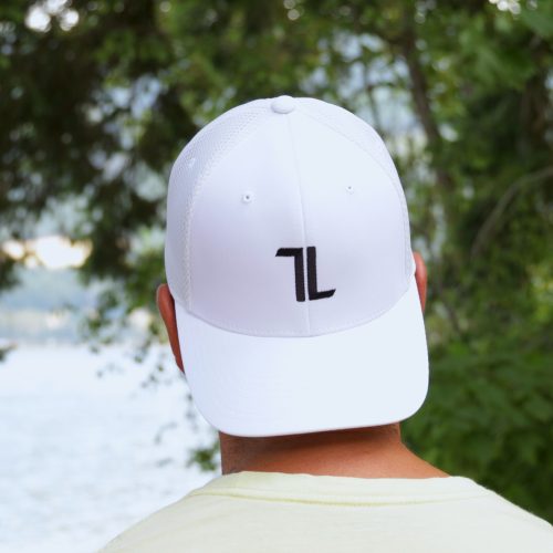 A person stands outdoors by water and green trees, wearing the Mesh Ball Cap - White with a minimalist flex fit, breathable mesh design, and black "7L" logo. Their face is not visible.