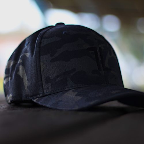 The Original Ball Cap - MultiCam, featuring a black and dark gray camouflage baseball cap design, sits on a wooden surface with a blurred outdoor background.