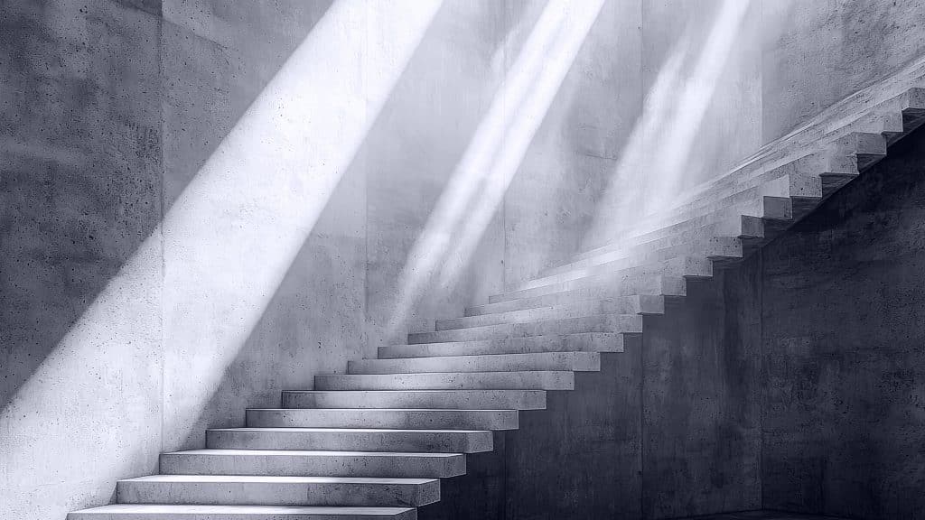 A minimalistic concrete staircase ascends alongside a blank wall, illuminated by dramatic beams of sunlight streaming in from above, creating sharp shadows and a serene, modern atmosphere.