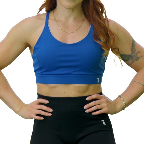 A woman with long red hair smiles with her hands on her hips, wearing the Everyday Ease Bra in blue and black leggings, standing against a white background.