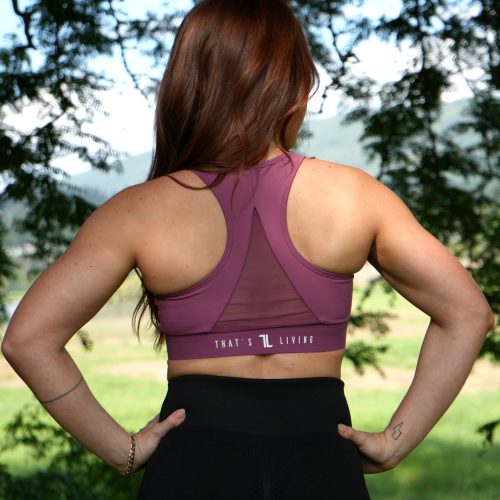 A woman with long brown hair stands outdoors with her back to the camera, wearing a ComfiZip Bra in pink and black leggings. Trees and a blurred landscape are visible in the background.