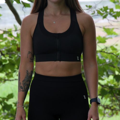 A woman with long brown hair, a left forearm tattoo, and a watch on her right wrist stands outdoors in front of green trees, wearing the ComfiZip Bra - Black and leggings.
