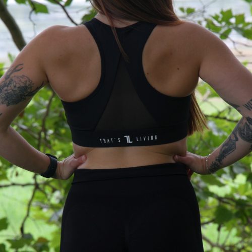 A woman with long hair, tattoos on both arms, stands outdoors with her back to the camera, wearing the ComfiZip Bra - Black and leggings. She has her hands on hips amid lush green foliage.