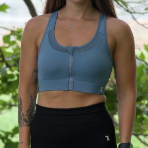 A woman with long brown hair stands outdoors in front of green trees, wearing the ComfiZip Bra in blue and black leggings. She has tattoos on her left arm, and accessorizes with a watch and necklace.