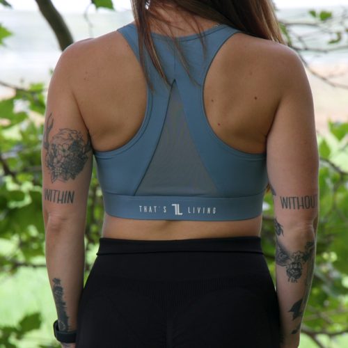 A woman with long brown hair stands outdoors, facing away. She wears a ComfiZip Bra - Blue and black leggings. Tattoos decorate both arms, including "WITHIN" on her left tricep and "WITHOUT" on her right tricep.