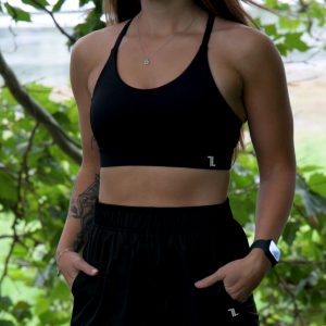 Wearing the Everyday Ease Bra - Black and shorts, a woman with long brown hair stands outdoors amid green foliage, hands in her pockets, and sports a fitness tracker on her wrist.