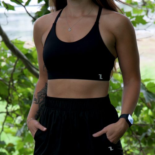 Wearing the Everyday Ease Bra - Black and shorts, a woman with long brown hair stands outdoors amid green foliage, hands in her pockets, and sports a fitness tracker on her wrist.
