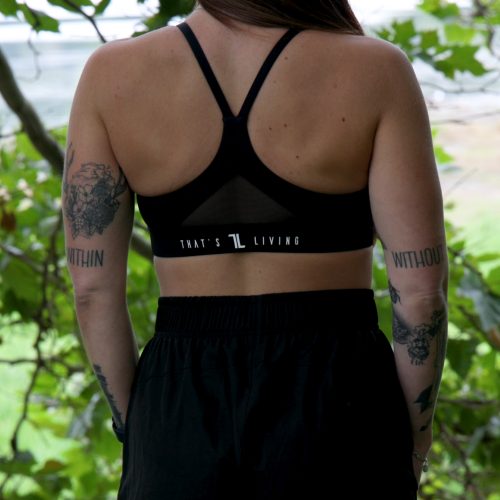 A woman with long brown hair and arm tattoos stands outdoors among greenery, wearing the Everyday Ease Bra - Black and black shorts, with her back to the camera.