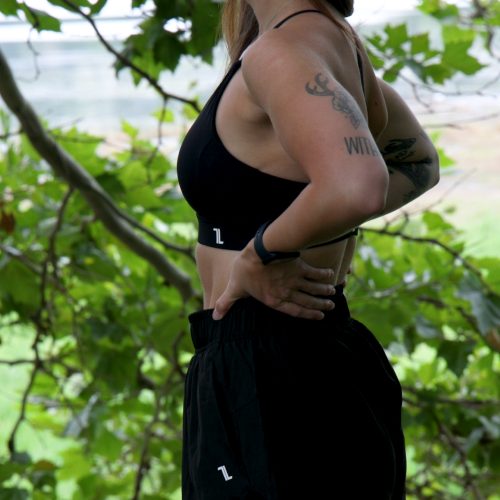 A woman wearing the Everyday Ease Bra in black and shorts stands outdoors, smiling with hands on her hips amid green leafy trees and a blurred natural background.