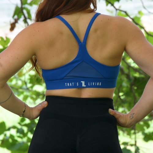 A woman with long auburn hair stands outdoors facing away, wearing the blue Everyday Ease Bra and black leggings, hands on her hips. Trees and greenery fill the background.