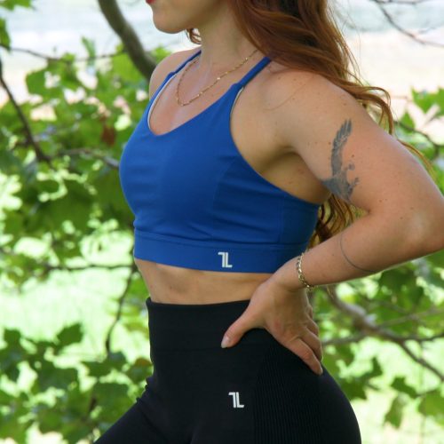 A woman with long red hair wears the Everyday Ease Bra in blue and black leggings outdoors, hands on hips, gazing sideways against a backdrop of green leaves and a blurred landscape.