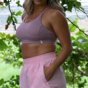 A woman with long blonde hair stands outdoors among green trees, wearing the Everyday Ease Bra in soft pink and light pink athletic shorts, looking at the camera with a serious expression.