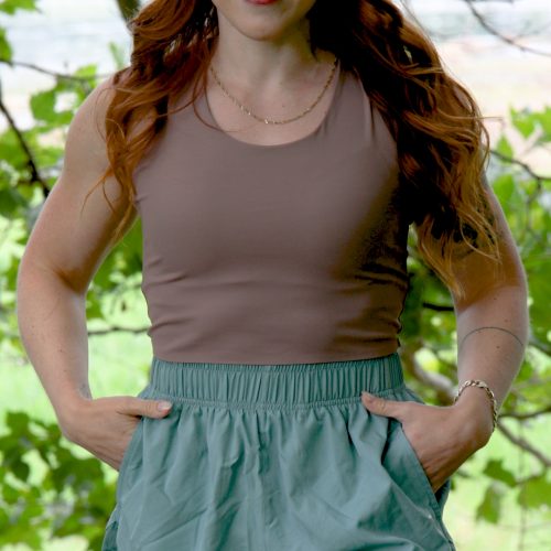 A woman with long red hair smiles outdoors, wearing a sleeveless taupe FlowEase BraTop - Mokarosa and light green shorts with her hands in the pockets. Green leaves and blurred scenery appear in the background.