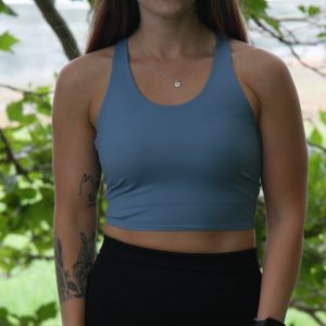A woman with long brown hair, wearing the FlowEase BraTop - Pastel Blue and black leggings, stands outdoors in front of leafy trees. She has tattoos on her left arm and accessorizes with a necklace and smartwatch.