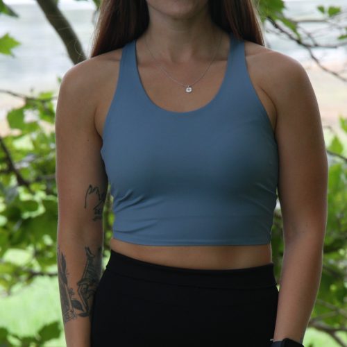 A woman with long brown hair, wearing the FlowEase BraTop - Pastel Blue and black leggings, stands outdoors in front of leafy trees. She has tattoos on her left arm and accessorizes with a necklace and smartwatch.