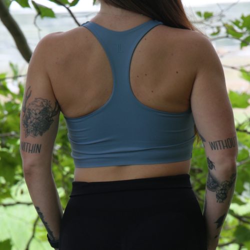 A woman with long brown hair wears a FlowEase BraTop - Pastel Blue and black leggings, standing outdoors with her back to the camera. Tattoos are visible on both arms, surrounded by lush green foliage.