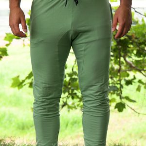 A person wearing Genesis Joggers - Fern Green, featuring an adjustable fit and zipper pockets, stands on grass with white socks. Leafy green trees and a blurred outdoor scene are in the background; their upper body is partially visible.