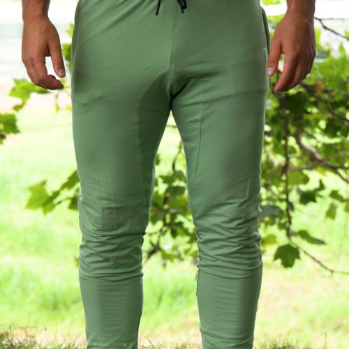 A person wearing Genesis Joggers - Fern Green, featuring an adjustable fit and zipper pockets, stands on grass with white socks. Leafy green trees and a blurred outdoor scene are in the background; their upper body is partially visible.