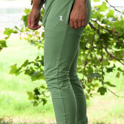 A person wears Genesis Joggers - Fern Green with adjustable fit and zipper pockets, paired with a black shirt and white shoes, standing on grass amid leafy green trees and a blurred outdoor background.