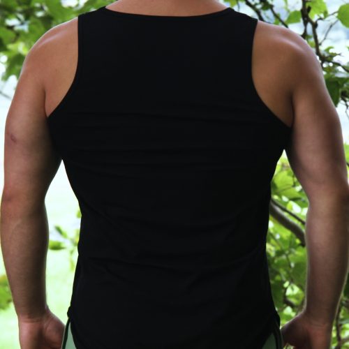 Wearing a black tank top and Genesis Joggers - Fern Green featuring an adjustable fit and zipper pockets, a person stands outdoors with their back to the camera, surrounded by leafy branches and greenery.
