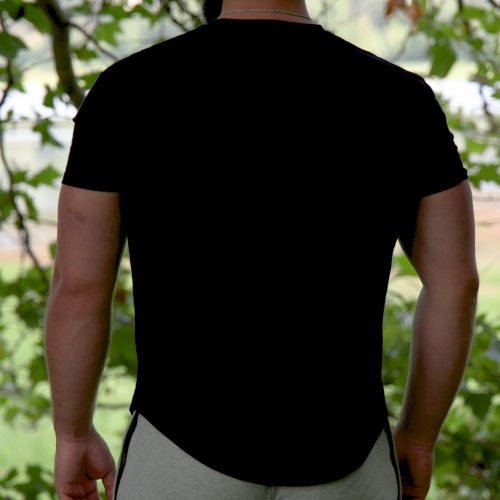 A bearded man stands outdoors, facing away from the camera, wearing the Genesis Tee - Black with a tapered fit and scoop bottom, paired with light gray shorts amid leafy green trees and a blurred background.