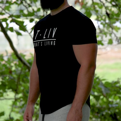 A bearded man stands outdoors among green trees, wearing the Genesis Tee - Black, a men’s performance t-shirt with tapered fit and scoop bottom, paired with light gray pants; grass and water appear blurred in the background.