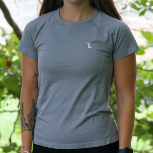 A woman with long brown hair stands outdoors before green foliage, wearing the GlideFlex Tee - Grey, black pants, a smartwatch, and showing tattoos on her left arm. She looks slightly to the side with a faint smile.