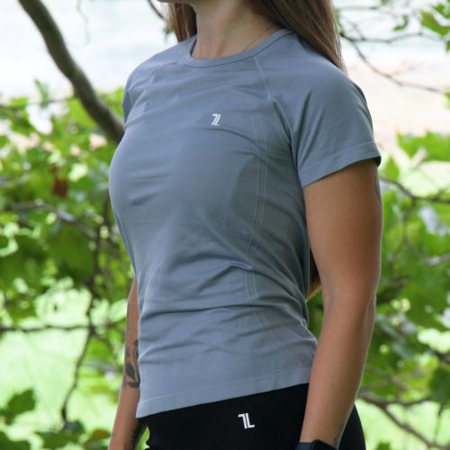 A woman with long brown hair stands outdoors among green leaves, wearing the GlideFlex Tee - Grey with black leggings. She is facing slightly left and wears a black watch on her wrist.