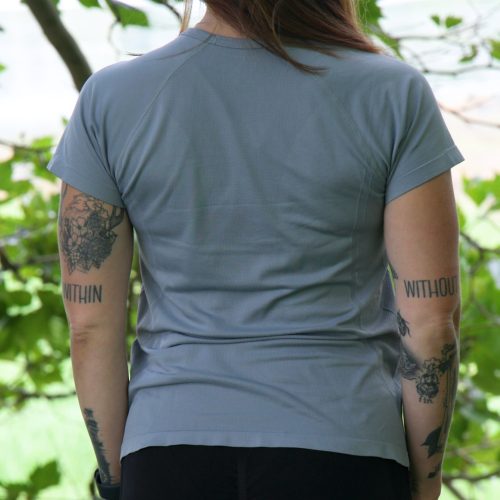 A person with long brown hair and tattoos on both arms, wearing the GlideFlex Tee - Grey and black pants, stands outdoors facing away amid green leafy trees.