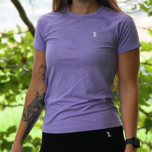 A woman with long brown hair stands outdoors before green foliage, smiling slightly. She wears a purple GlideFlex Tee, black leggings, has a tattoo on her left arm, and a black wristwatch on her right wrist.