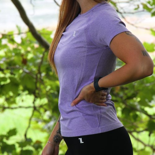 A woman with long hair stands outdoors in profile, wearing the GlideFlex Tee - Purple and black leggings, one hand on her hip amid lush green foliage.