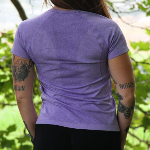 A person with long brown hair and tattoos on both arms, wearing the GlideFlex Tee - Purple and black pants, stands outdoors facing away, surrounded by green leaves and trees.