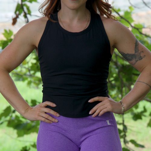 A red-haired woman wears the LuxeForm Tank - Black while standing outdoors with her hands on her hips, smiling confidently. Green leaves and trees form the background.