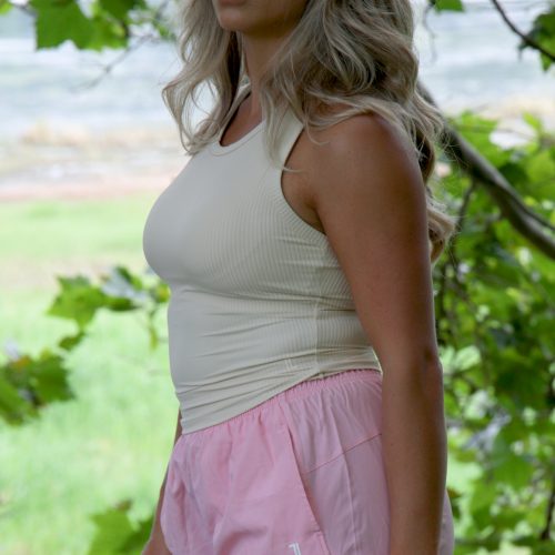 A woman with long, wavy blonde hair stands outside among green trees, wearing the LuxeForm Tank - Cream White and light pink shorts, gazing ahead with a neutral expression.